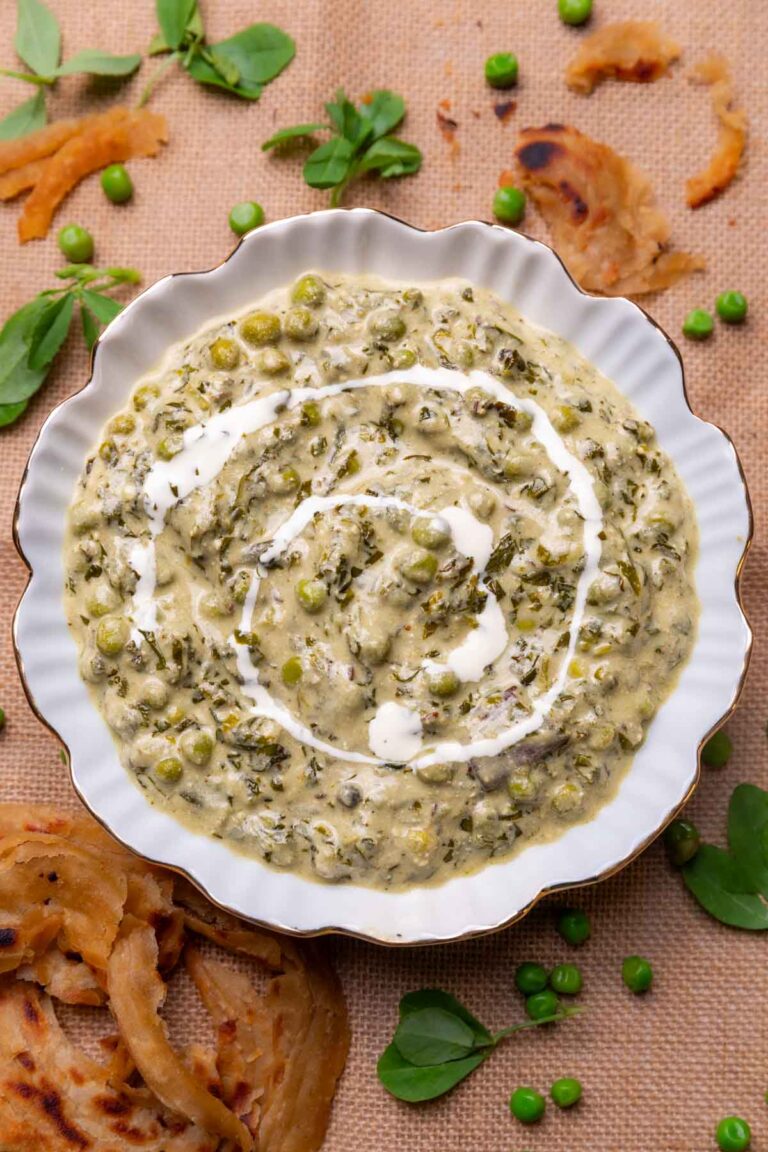 Methi Matar Malai served in a white bowl with lacha paratha, fenugreek leaves, matar etc strewn about